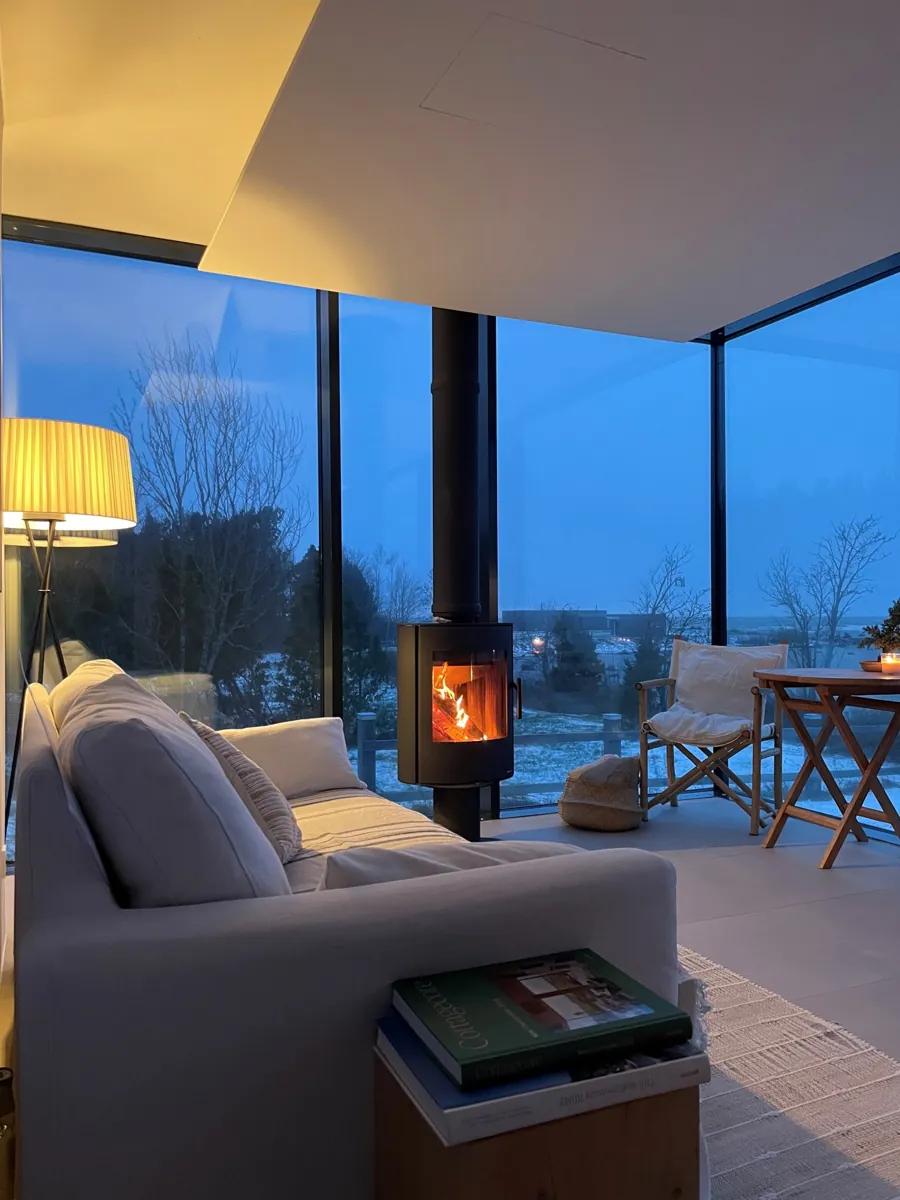 Inside a Signature in winter: a wood stove burning, a sofa and a lamp, snow beyond the glass