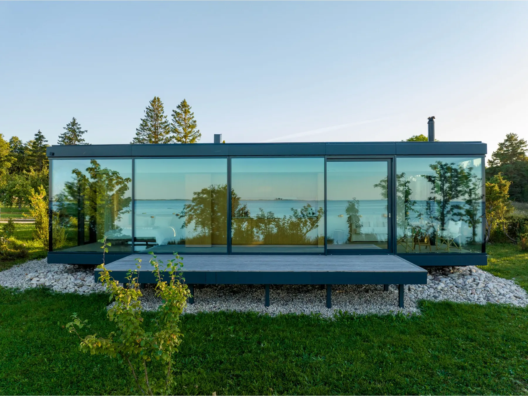 Etnika Signature glass house on a lawn above the sea, the landscape reflected in its long facade