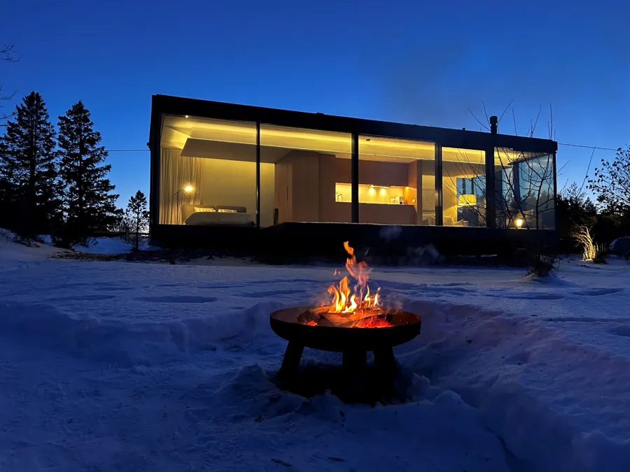 Signature glass house lit at dusk in snow, a fire bowl burning in front of it